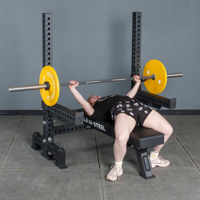 A person in athletic clothing prepares to bench press with a barbell and yellow plates on the Bells of Steel Manticore Two Post Converter Bench in a gym with gray walls and a concrete floor.