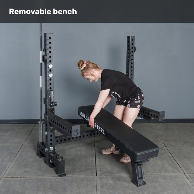 Wearing athletic gear, a woman adjusts the Manticore Two Post Converter Bench by Bells of Steel on a black steel rack. The minimalist gym features gray walls and floor. "Removable bench" is displayed above.