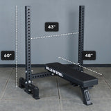 A black Manticore Two Post Converter Bench by Bells of Steel stands at a bench station on a gray floor, with vertical supports 60" high, 43" wide, and the bench measuring 48" long.