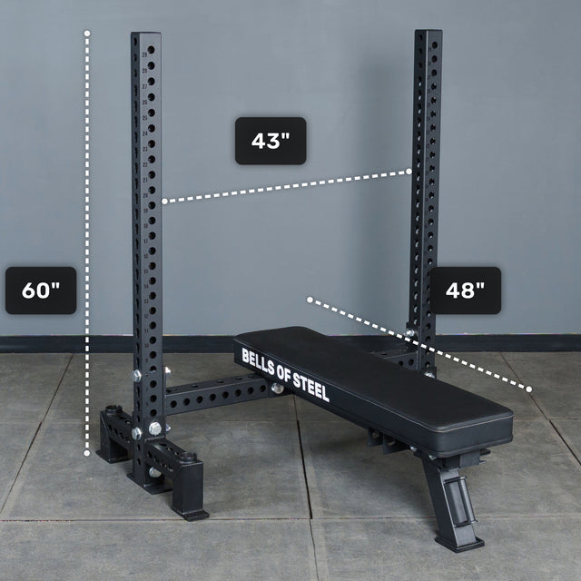 A black Manticore Two Post Converter Bench by Bells of Steel stands at a bench station on a gray floor, with vertical supports 60" high, 43" wide, and the bench measuring 48" long.