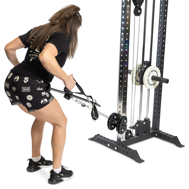 A person with long hair, dressed in a black T-shirt and shorts, performs a cable row exercise using the 30 D-Handle Bar Cable Attachment with Carabiners. This product from Bells of Steel is pulled towards their waist while they are slightly bent over, engaging their back muscles.