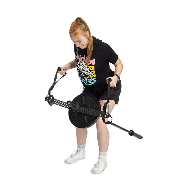 A red-haired individual, dressed in a black T-shirt and shorts, is using the Bells of Steel 30 D-Handle Bar Cable Attachment with Carabiners to perform resistance exercises on a portable pulley gym. They remain in a bent-over position while holding the device's handles.