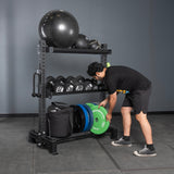A man in black athletic wear places a green weight plate onto the Bells of Steel Storage Shelves, which also hold medicine balls, dumbbells, and other fitness equipment against a gray wall.