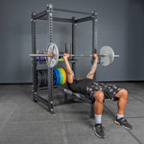 A person bench presses a barbell on a weight bench inside a power rack, while Bells of Steel Storage Shelves on the rack hold weight plates and gym equipment in a gym with gray walls and flooring.