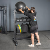 A man in athletic wear places a large black exercise ball on the top Bells of Steel Storage Shelves, which hold weights, bags, and other fitness equipment against a gray wall.