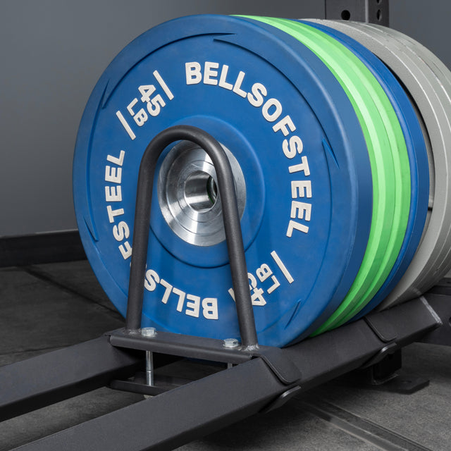 A close-up shows Bells of Steel weight plates loaded on a barbell, with blue, green, and gray plates in a gym featuring Storage Shelves in the background.