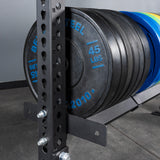 A close-up of Bells of Steel Storage Shelves displays several 45-pound black weight plates on a gym weight rack, with a metal support post featuring numbered holes; the gym floor and gray wall appear in the background.