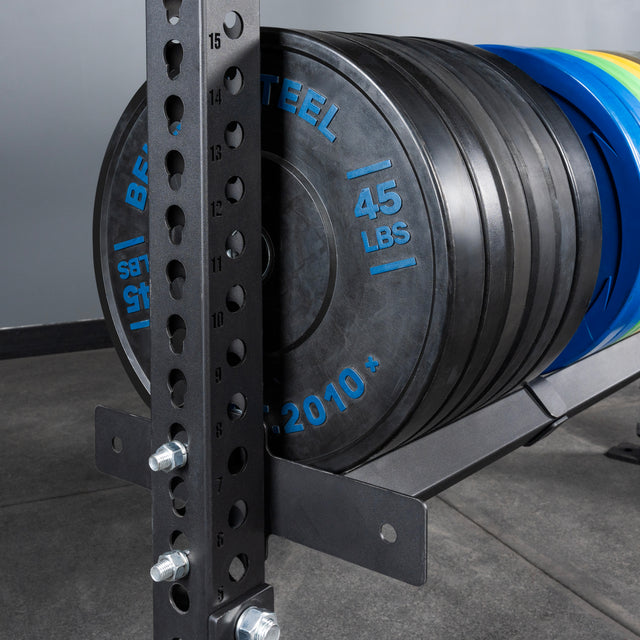 A close-up of Bells of Steel Storage Shelves displays several 45-pound black weight plates on a gym weight rack, with a metal support post featuring numbered holes; the gym floor and gray wall appear in the background.