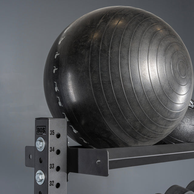 A large black exercise stability ball rests on a Bells of Steel Storage Shelf against a gray backdrop. The ball features circular textured lines with partial white numbers and symbols visible.