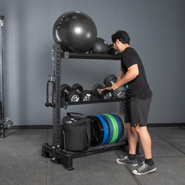 Wearing athletic clothes, a person picks up a dumbbell from Bells of Steel Storage Shelves, which hold exercise equipment like medicine balls, weight plates, and sandbags in a gym with gray walls and flooring.