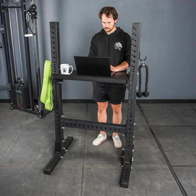 A man uses a laptop on Bells of Steel Storage Shelves in a gym. A green resistance band, white mug, and workout rope hang from the shelves. The gray floor and other gym equipment are visible in the background.