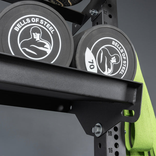 Close-up of two black Bells of Steel weight plates with white logos on a Bells of Steel Storage Shelf, next to a green resistance band hanging from the side.