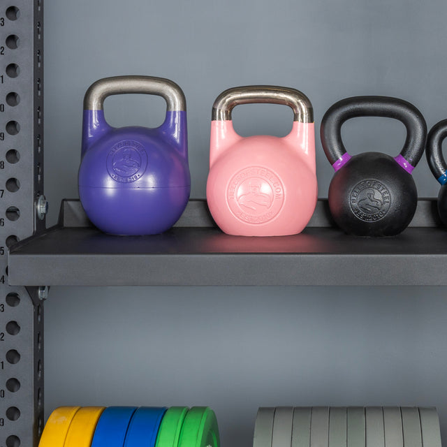 Three kettlebells rest on the Bells of Steel Storage Shelves, with colorful weight plates organized below. The shelf and background wall are gray.