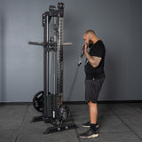A man in black athletic wear prepares to do a cable bicep curl with the Bells of Steel Oblivyon Tower - 3" x 3” Cable Tower in a gym, with gray walls, flooring, and a squat rack seen in the background.