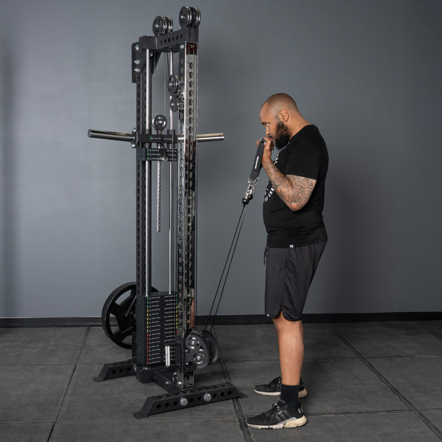 A man in black athletic wear prepares to do a cable bicep curl with the Bells of Steel Oblivyon Tower - 3" x 3” Cable Tower in a gym, with gray walls, flooring, and a squat rack seen in the background.