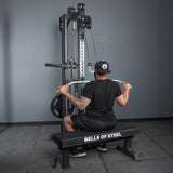 A person in black athletic wear performs a lat pulldown on a Bells of Steel Oblivyon Tower - 3" x 3” Cable Tower while seated on a bench labeled “BELLS OF STEEL” in a gym with gray walls and flooring.