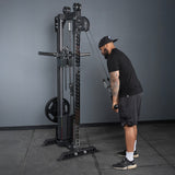 Wearing black workout clothes, a person uses the Bells of Steel Oblivyon Tower - 3" x 3” Cable Tower for tricep exercises in a gym with gray walls, rubber flooring, and a squat rack in the background.