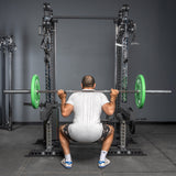 A man in a white t-shirt and shorts performs a squat with a barbell loaded with green plates on the Bells of Steel Oblivyon Tower 3" x 3” Cable Tower Squat Stands, near adjustable cable crossover machines. The image is viewed from behind.