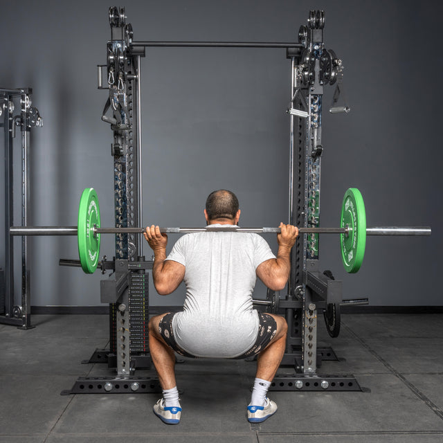 A man in a white t-shirt and shorts performs a squat with a barbell loaded with green plates on the Bells of Steel Oblivyon Tower 3" x 3” Cable Tower Squat Stands, near adjustable cable crossover machines. The image is viewed from behind.