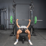 A person bench presses a barbell with green plates inside a squat rack, wearing a white shirt, black shorts, and white sneakers. Nearby are Bells of Steel Oblivyon Tower - 3" x 3" Cable Tower Squat Stands.