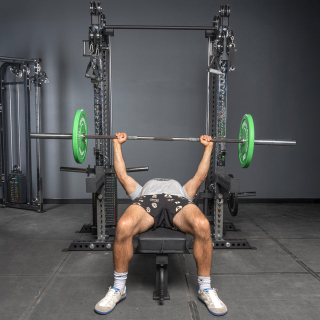 A person bench presses a barbell with green plates inside a squat rack, wearing a white shirt, black shorts, and white sneakers. Nearby are Bells of Steel Oblivyon Tower - 3" x 3" Cable Tower Squat Stands.