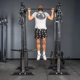 A man in a t-shirt, printed shorts, and sneakers does pull-ups on a Bells of Steel Oblivyon Tower - 3" x 3” Cable Tower Squat Stand in a gym with grey walls and a concrete floor.
