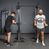 Two men work out at a gym with gray walls and black flooring. One uses the Bells of Steel Oblivyon Tower - 3" x 3” Cable Tower Squat Stands with a rope attachment, while the other stands nearby curling a bar with both hands.