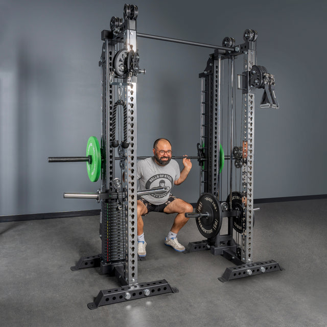 A man in a gray t-shirt and shorts performs a squat with a barbell on Bells of Steel’s Oblivyon Tower - 3" x 3” Cable Tower Squat Stands, featuring weights, pulleys, and an adjustable cable crossover. A plain gray wall is in the background.