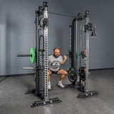 A man wearing a gray T-shirt squats with a barbell inside the Bells of Steel Oblivyon Tower 3" x 3” Cable Tower Squat Stands, in a room with gray walls and floor.
