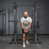 A bearded man with glasses uses the Bells of Steel Oblivyon Tower – 3" x 3” Cable Tower Squat Stands for a chest workout, wearing a white “Build4Steel” t-shirt, black shorts, and white sneakers in a gray-walled gym.