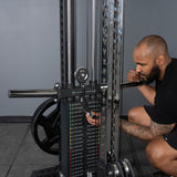 A bearded man with a tattooed right arm, dressed in a black shirt and shorts, kneels beside the Bells of Steel Oblivyon Tower - 3" x 3” Cable Tower Squat Stands, adjusting the weight pin in a gym.