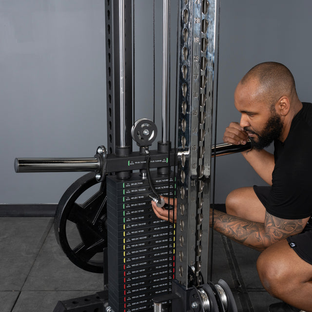 A bearded man with a tattooed right arm, dressed in a black shirt and shorts, kneels beside the Bells of Steel Oblivyon Tower - 3" x 3” Cable Tower Squat Stands, adjusting the weight pin in a gym.