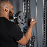 A man in a black t-shirt adjusts the pulley on the Bells of Steel Oblivyon Tower - 3" x 3” Cable Tower Squat Stands in a gym setting.