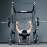 A person lies on a bench inside the Bells of Steel Manticore Four Post Power Rack Builder (3" x 3", 1" Holes), gripping a barbell loaded with weight plates above their chest as if preparing to perform a bench press.
