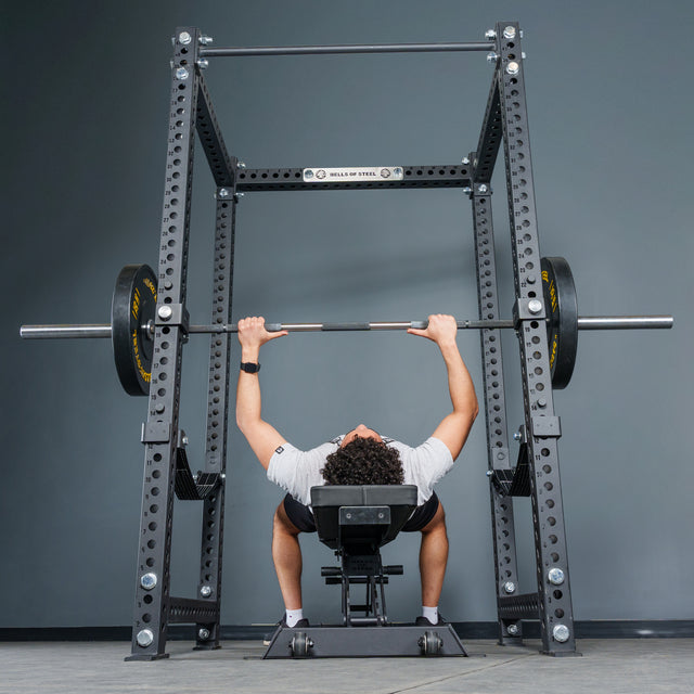 A person lies on a bench press, gripping a barbell loaded with weights, preparing to lift inside the Bells of Steel Manticore Four Post Power Rack – Prebuilt (3" x 3", 1" Holes) positioned against a gray wall.