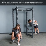 Two people use the Bells of Steel Manticore Four Post Power Rack – Prebuilt (3" x 3", 1" Holes) with attachments; one does lunges holding dumbbells, while the other performs a bent-over row with a barbell. Text above reads: “Rack attachments unlock even more workouts—perfect for building your home gym.”.