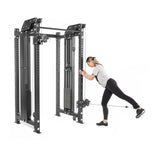 A woman in sports attire demonstrates a leg exercise with the Kraken Rack Attachment for 4 Post Hydra & Manticore Racks by Bells of Steel. She balances on one leg, extending the other back while holding onto the sleek black equipment, which features multiple pulleys for an efficient cable system.