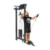 A man in a black T-shirt and shorts exercises using the Bells of Steel Kraken Rack Attachment for 4 Post Hydra & Manticore Racks (Single Side). He stands on a platform, holding cables, and extends his arms outward. The machine allows precise adjustments with its stacked weight system.