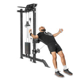 A bearded, bespectacled person is working out on a Bells of Steel Kraken Rack Attachment for 4 Post Hydra & Manticore Racks (Single Side). They're in a black T-shirt, black shorts, white socks, and athletic shoes, stretching backward with the machine's cables.