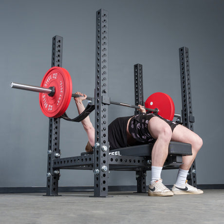 A person uses the Bells of Steel Hydra Four Post Converter Bench in a gym, lifting a barbell with red plates. They wear a black shirt, shorts, white socks, beige sneakers, and have resistance bands around their thighs.