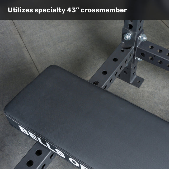 A close-up of the Bells of Steel Hydra Four Post Converter Bench with a specialty 43-inch crossmember and large bolts on a gray floor. The floating design boosts powerlifting stability. Text reads, "Utilizes specialty 43” crossmember.