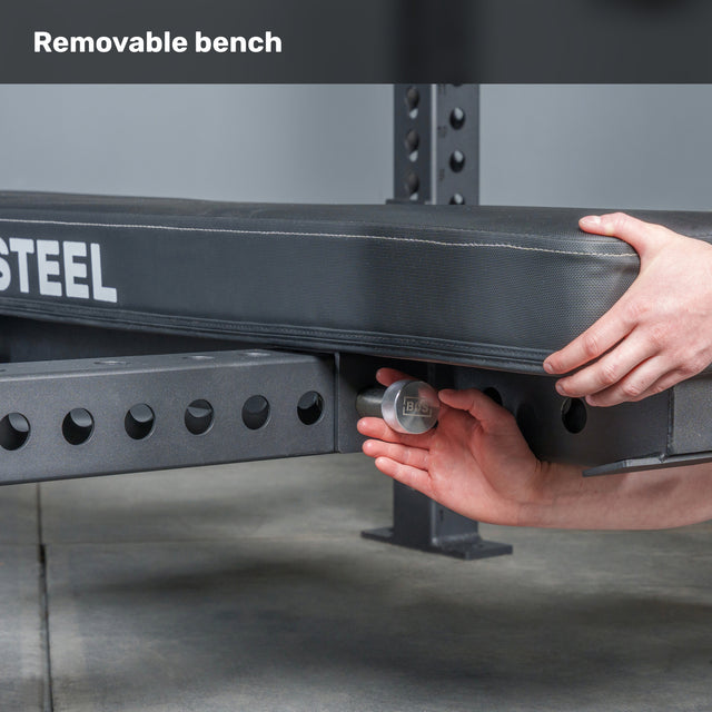 A person adjusts the Manticore Four Post Converter Bench by Bells of Steel, turning a knob under its padded seat on a metal frame with multiple holes. The word "STEEL" is nearby, with "Removable bench" at the top.