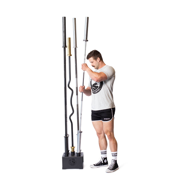 A person in a gray T-shirt, black shorts, and black shoes stands beside the Bells of Steel 5 Barbell Holder, crafted from solid steel for secure organization of three barbell types, highlighting smart storage.