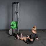 A woman performs a seated row on a Bells of Steel Footplate for Cable Tower/All In One Trainer with green weight plates in a minimalist gym with gray walls.