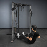 A person in black workout clothes sits on the floor, leaning back with arms behind them, facing a cable machine with a Bells of Steel Footplate for Cable Tower/All In One Trainer in a gym with gray walls and black flooring.