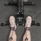 Wearing pink sneakers and patterned socks, a person sits at a rowing machine with feet on a Bells of Steel Footplate for Cable Tower/All In One Trainer, hands gripping the handles, poised to exercise atop gray gym mats.