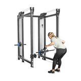 A person with a ponytail uses the Bells of Steel Kraken Rack Attachment for 6 Post Hydra Racks in the gym. They're wearing a gray shirt and black leggings, bent over and focused on strength training. The machine's metal frame and adjustable trolleys enhance their workout.