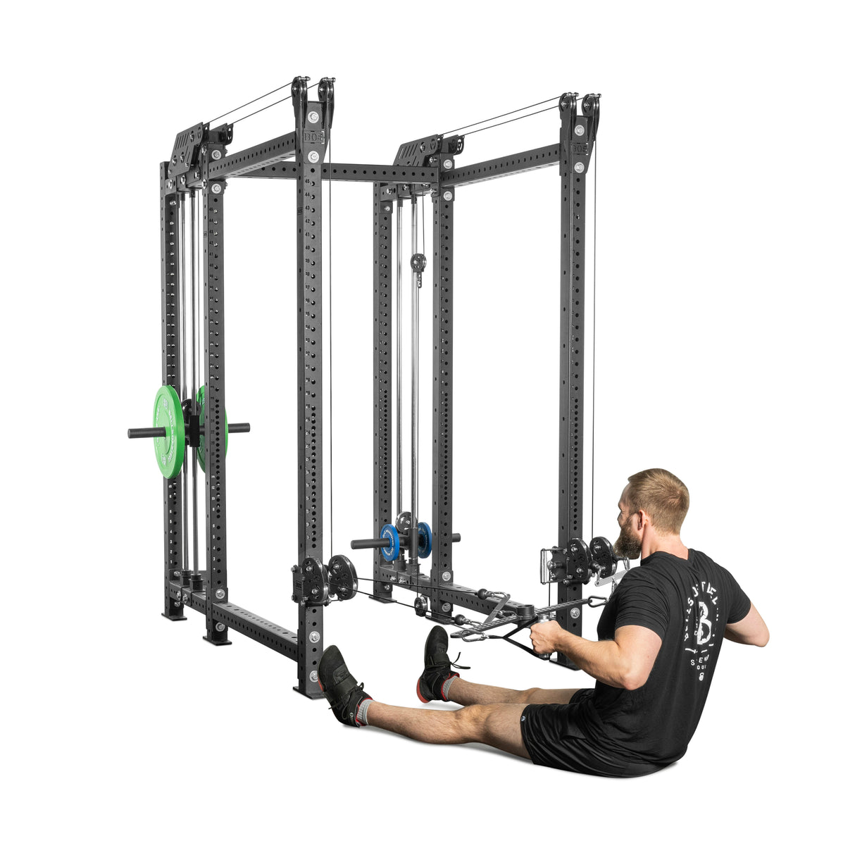 A person in a black t-shirt and shorts uses the Kraken Rack Attachment by Bells of Steel on a 6 Post Hydra Rack for seated rows, pulling the cable handles with both hands while seated on the floor. The large metal gym equipment has adjustable weights.