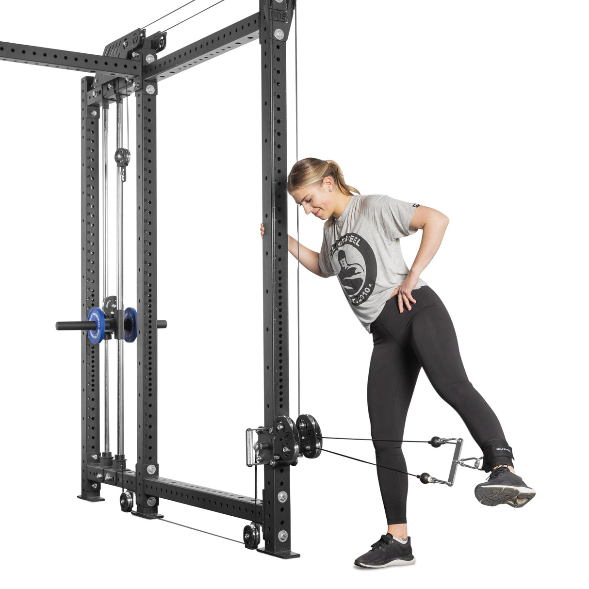 A woman in a gray t-shirt and black leggings performs a leg exercise using the Bells of Steel Kraken Rack Attachment for 6 Post Hydra Racks. She stands on one leg, holding the frame, while extending her other leg back with an adjustable trolley and strap around her ankle.