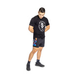 A person stands against a white background wearing a black Hollister t-shirt, Bells of Steel lightweight gym shorts with colorful patches made from stretchy high-performance polyester, and black socks and shoes, looking downward with their hands behind their back.